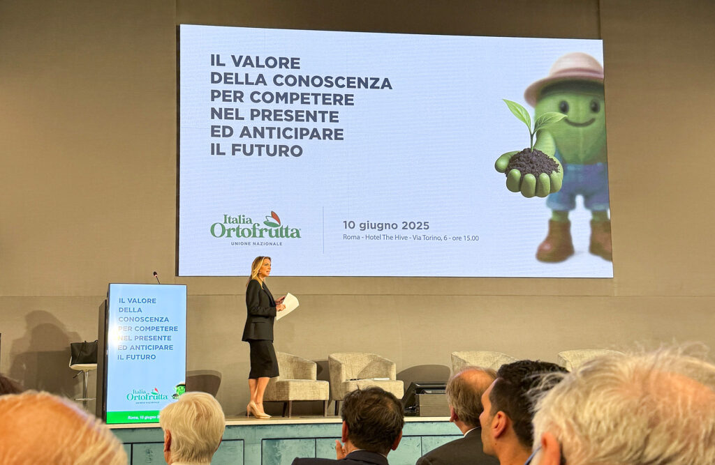 Il valore della filiera nel progetto di comunicazione integrata - Immagine 8 Scatto dal palco del convegno con la presentatrice e, sullo sfondo, la presentazione con il titolo dell’evento e la mascotte antropomorfa a forma di foglia.
